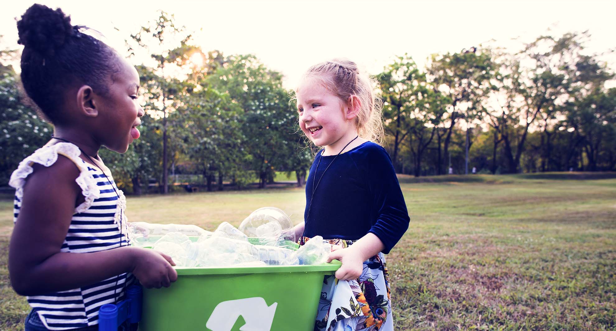 Two children, carry a recycling box in a garden.