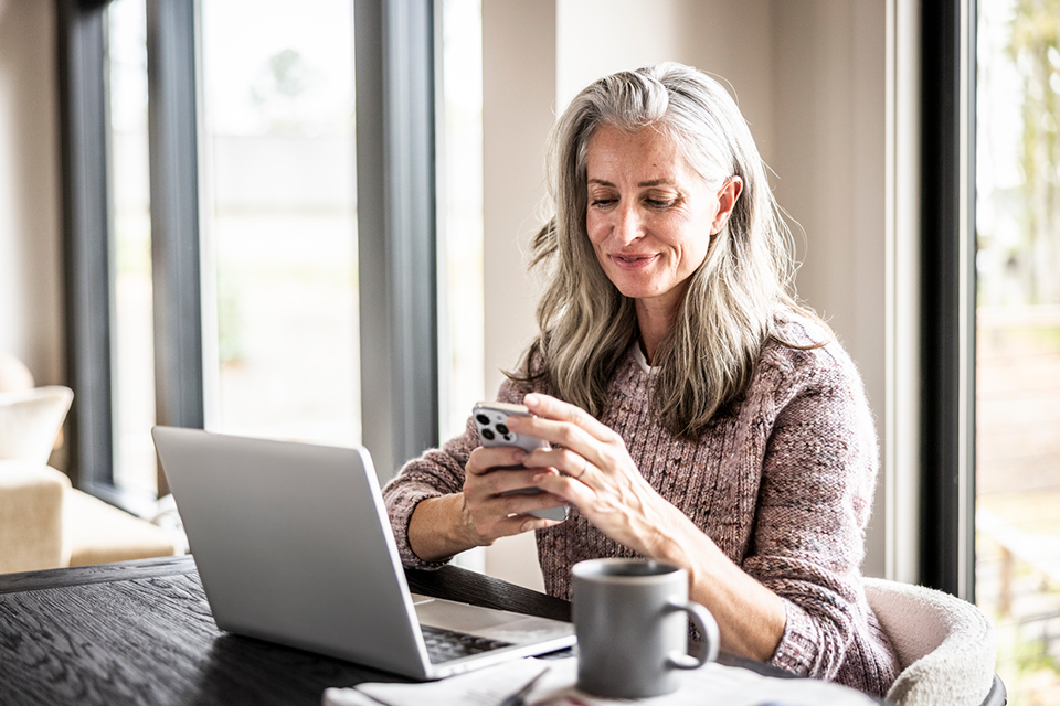A woman holding her phone while sitting at a desk with a cup and a laptop in front of her, she is smiling at her phone
