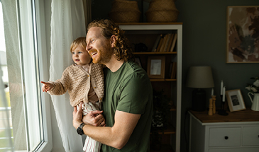 A man is holding his daughter, they are looking out a window in their home