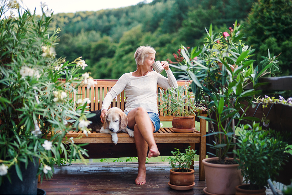 A woman is sitting on a bench in her garden, surrounded by plants. She is sipping from a cup and is petting her dog, who is lying beside her
