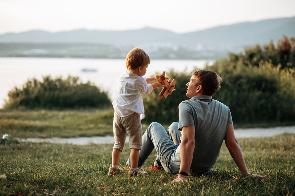 A father sitting in the grass while his son stands next to him, playing with a toy plane